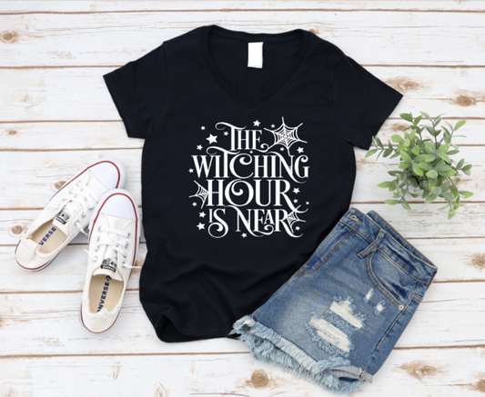 Black t-shirt with 'The Witching Hour is Near' text, white sneakers, and denim shorts on a wooden surface. www.missmafia.com