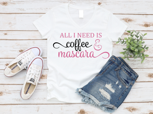 White V-neck t-shirt with the phrase "All I Need Is Coffee & Mascara" in white and pink text, styled with white Converse sneakers, ripped denim shorts, and a green plant on a white wooden background.
