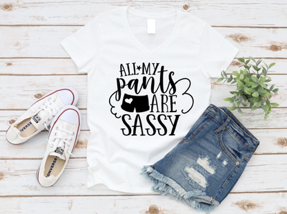 White V-neck t-shirt with the phrase "All My Pants Are Sassy" in bold black lettering, styled with distressed denim shorts, white Converse sneakers, and a small potted plant on a whitewashed wooden background.