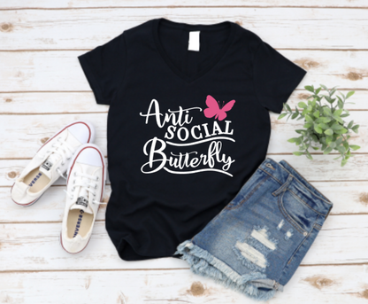 Black V-neck t-shirt with “Anti Social Butterfly” text and a pink butterfly graphic, styled alongside distressed denim shorts, white Converse sneakers, and a potted plant on a whitewashed wooden backdrop.