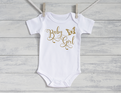 White baby onesie with 'Baby Girl' and butterfly design in gold on a wooden background