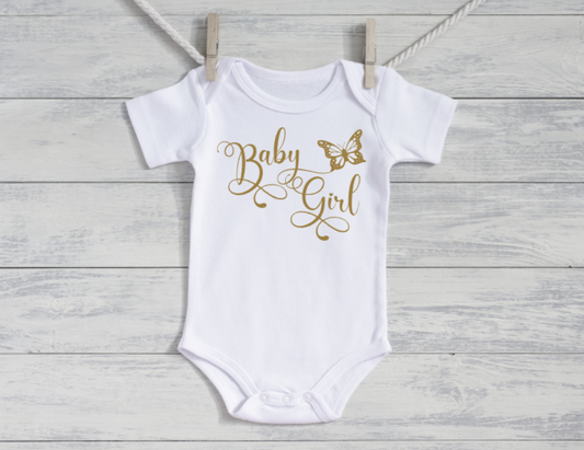 White baby onesie with 'Baby Girl' and butterfly design in gold on a wooden background