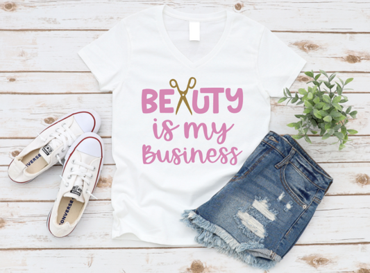 White t-shirt with pink “Beauty is my Business” text and gold scissors graphic, styled with white sneakers and distressed denim shorts on a wooden background.