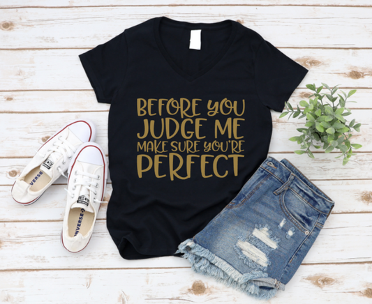Black t-shirt with gold text reading “Before you judge me make sure you’re perfect,” paired with denim shorts and white sneakers on a light wooden background.