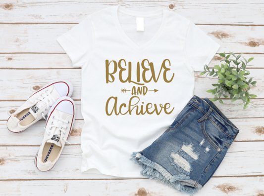 White t-shirt with “BELIEVE AND ACHIEVE” text in gold, styled with white sneakers and ripped denim shorts on a wooden background.