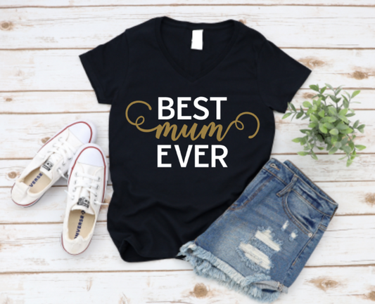 Black t-shirt with "Best Mum Ever" text in black and gold, styled with white sneakers and ripped denim shorts on a wooden background.