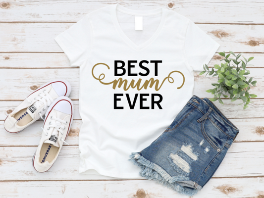 White t-shirt with "Best Mum Ever" text in black and gold, styled with white sneakers and ripped denim shorts on a wooden background.