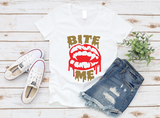 White t-shirt with bold red vampire lips and dripping "Bite Me" text, styled with white sneakers and ripped denim shorts on a light wooden background.