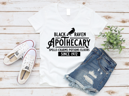 White t-shirt with “Black Raven Apothecary – Spells, Charms, Potions, Elixirs – Since 1972” printed in white. Styled with ripped denim shorts, white sneakers, and greenery on a white wooden backdrop.