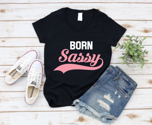 Black t-shirt with "Born Sassy" pink retro text, styled with white sneakers and ripped denim shorts on a white wood background.