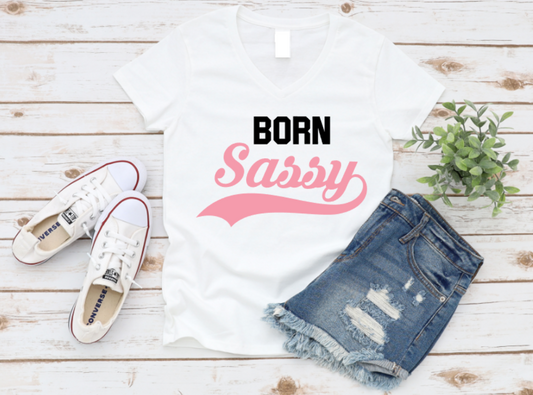 White t-shirt with "Born Sassy" pink retro text, styled with white sneakers and ripped denim shorts on a white wood background.