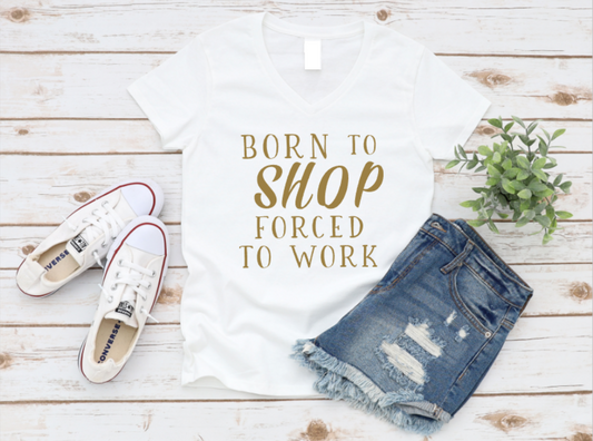 White t-shirt with "Born to Shop Forced to Work" styled with white sneakers and distressed denim shorts on a white wood background.