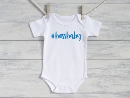 White baby onesie with '#bossbaby' text on a wooden surface