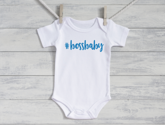 White baby onesie with '#bossbaby' text on a wooden surface