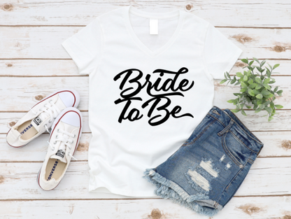 White t-shirt with "Bride To Be" in bold black script, styled with ripped denim shorts and white sneakers on a white wooden background.