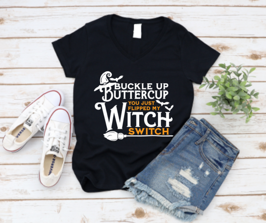 Black t-shirt with "Buckle Up Buttercup – You Just Flipped My Witch Switch" text in black and orange, styled with ripped denim shorts and white sneakers on a white wood background.