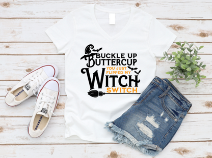 White t-shirt with "Buckle Up Buttercup – You Just Flipped My Witch Switch" text in black and orange, styled with ripped denim shorts and white sneakers on a white wood background.