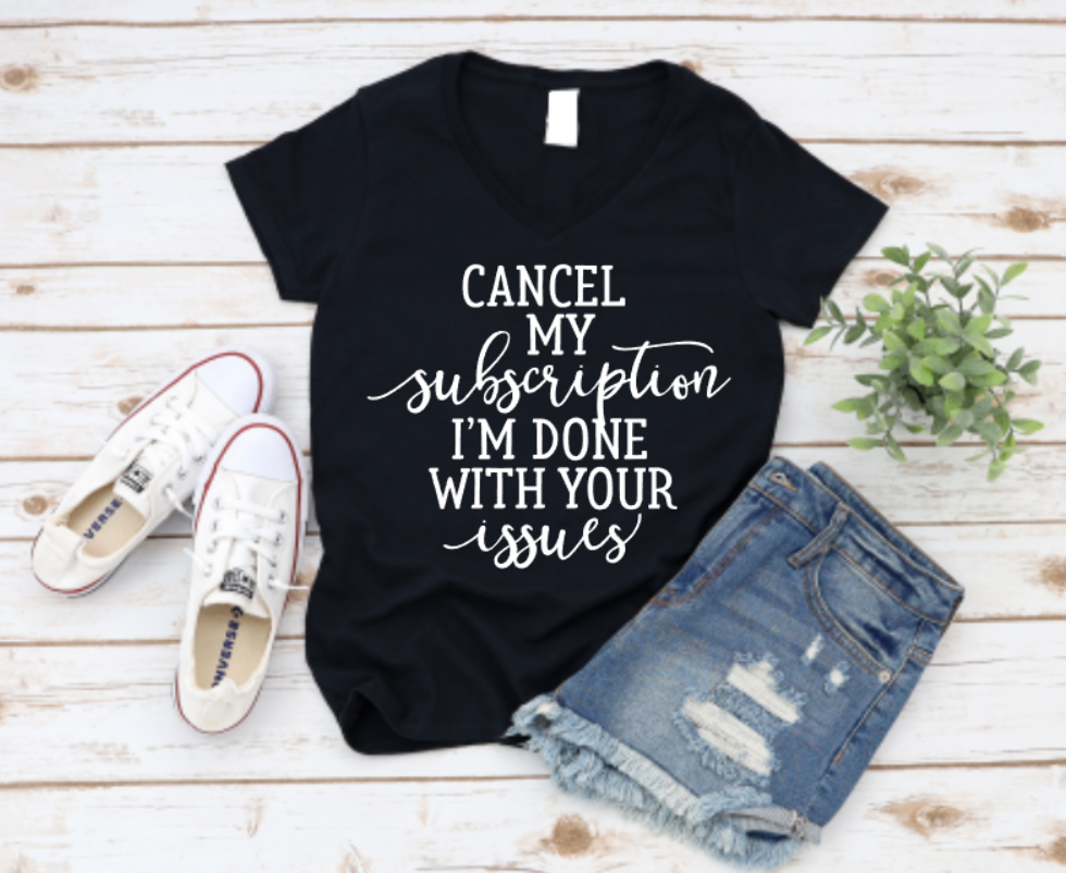 Black t-shirt with white “Cancel my subscription I’m done with your issues” text, styled with distressed denim shorts, white sneakers, and greenery on a wooden background.