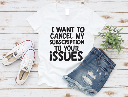 White t-shirt with the text “I want to cancel my subscription to your issues,” styled with ripped denim shorts, white sneakers, and a green plant on a wooden background.
