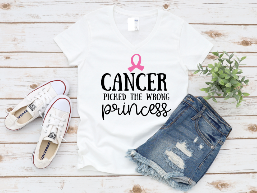 White t-shirt with “Cancer Picked the Wrong Princess” text and pink ribbon, styled with denim shorts and white sneakers on a wooden surface.