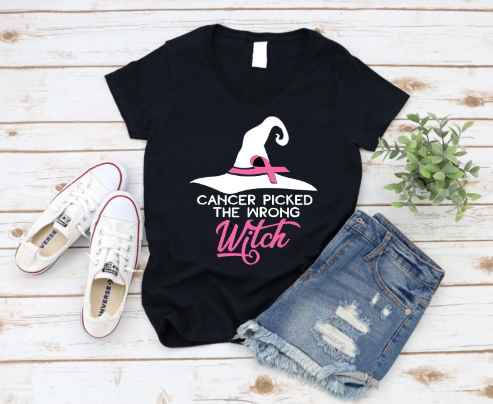 Black t-shirt with the phrase "Cancer Picked the Wrong Witch" in white and pink lettering, featuring a witch hat and pink ribbon design. Styled with denim shorts and white sneakers on a wooden background.