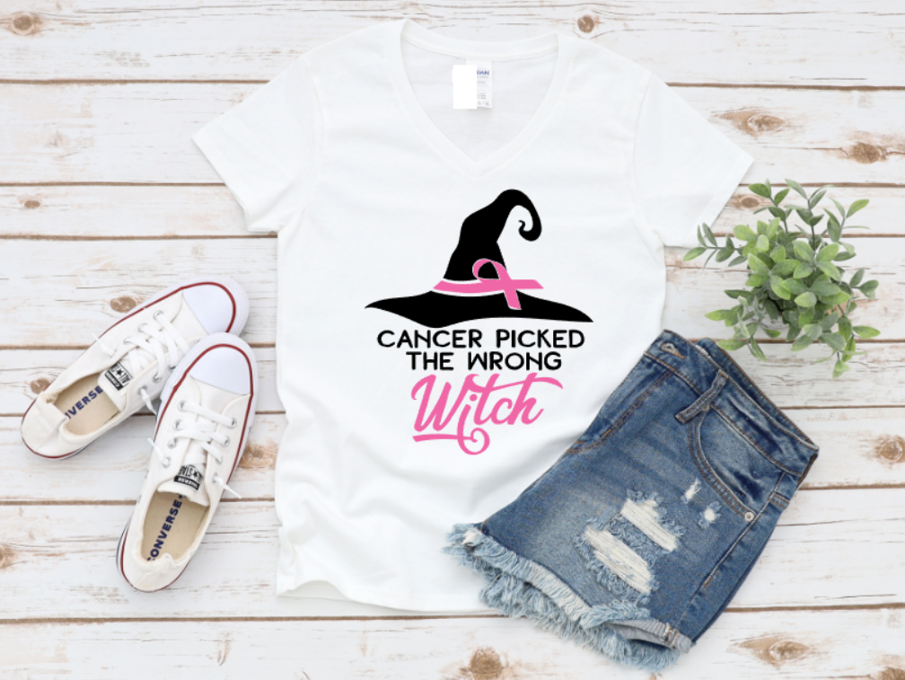 White t-shirt with the phrase "Cancer Picked the Wrong Witch" in white and pink lettering, featuring a witch hat and pink ribbon design. Styled with denim shorts and white sneakers on a wooden background.