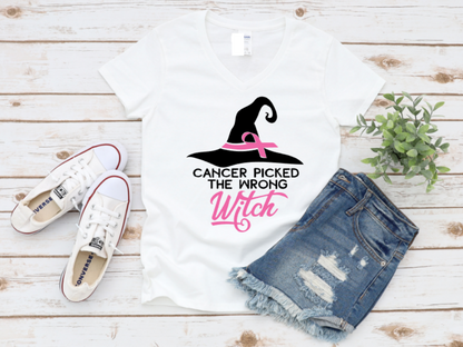 White t-shirt with the phrase "Cancer Picked the Wrong Witch" in white and pink lettering, featuring a witch hat and pink ribbon design. Styled with denim shorts and white sneakers on a wooden background.