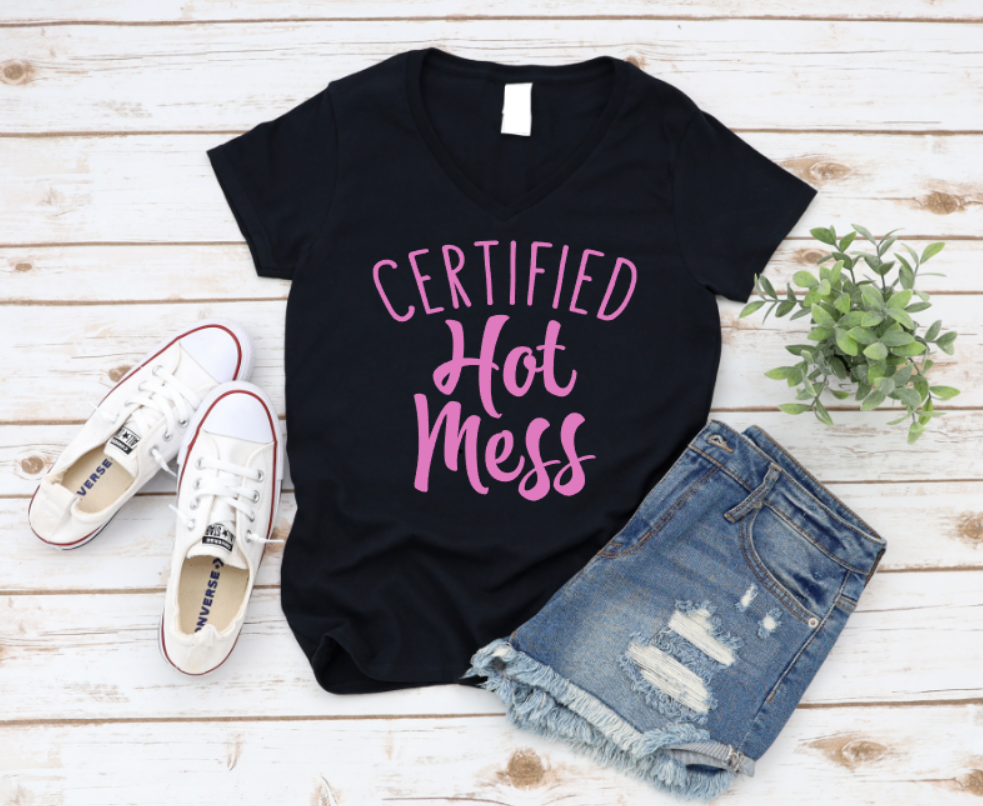 Black t-shirt with "Certified Hot Mess" text in pink, styled with white sneakers and distressed denim shorts on a wooden background.