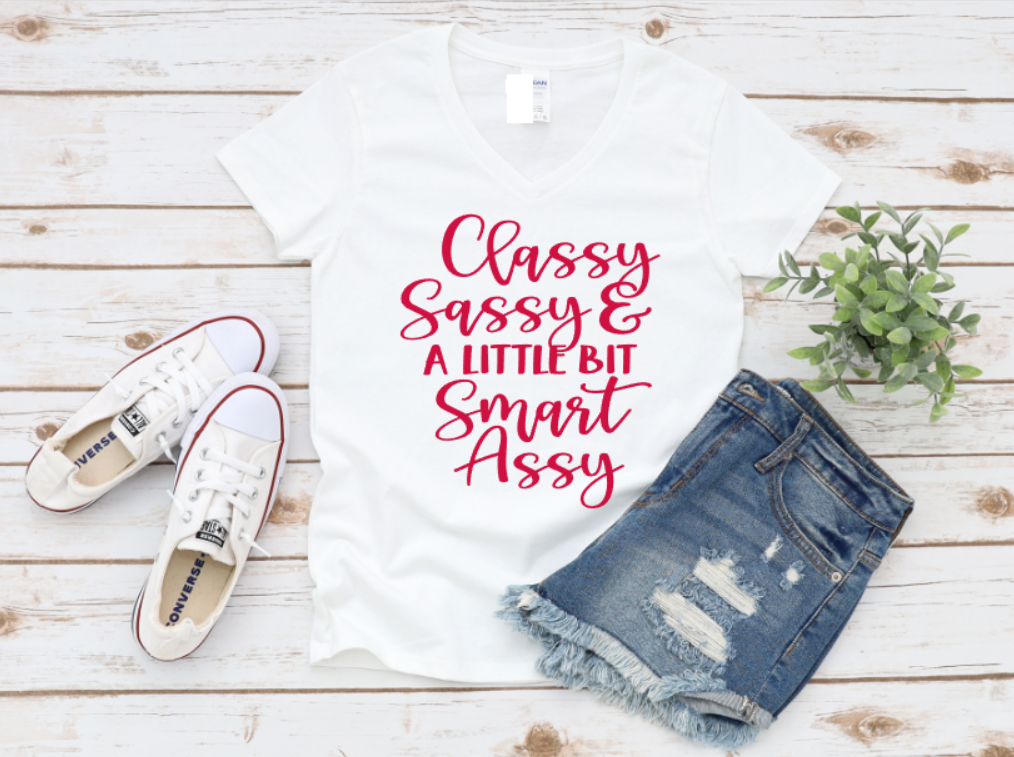 White t-shirt with “Classy Sassy & a Little Bit Smart Assy” red text, styled with denim shorts and white sneakers on a light wooden surface.