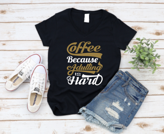 Black t-shirt with "Coffee Because Adulting Is Hard" design in brown, gold, and black lettering, styled with ripped denim shorts and white Converse sneakers on a wooden background.