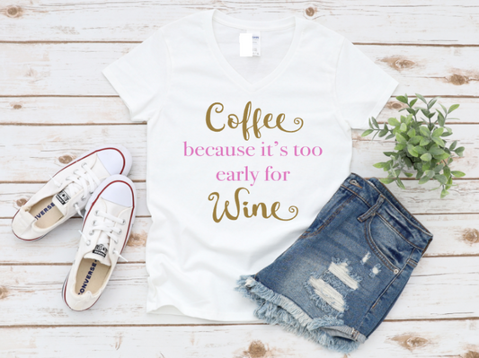 White t-shirt with the phrase "Coffee because it’s too early for Wine" in gold and pink text, styled with denim shorts, white sneakers, and a plant on a wooden surface.