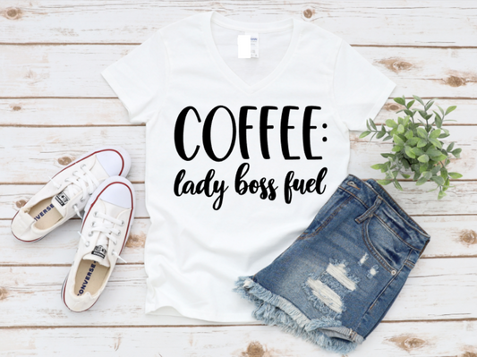 White t-shirt with bold black “Coffee: lady boss fuel” text, styled with ripped denim shorts and white Converse sneakers on a white wooden background.