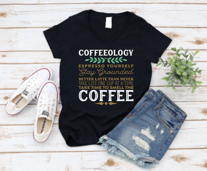 Black t-shirt with “Coffeeology” espresso-themed text in green, gold, and white, paired with distressed denim shorts and white Converse sneakers on a wooden background.
