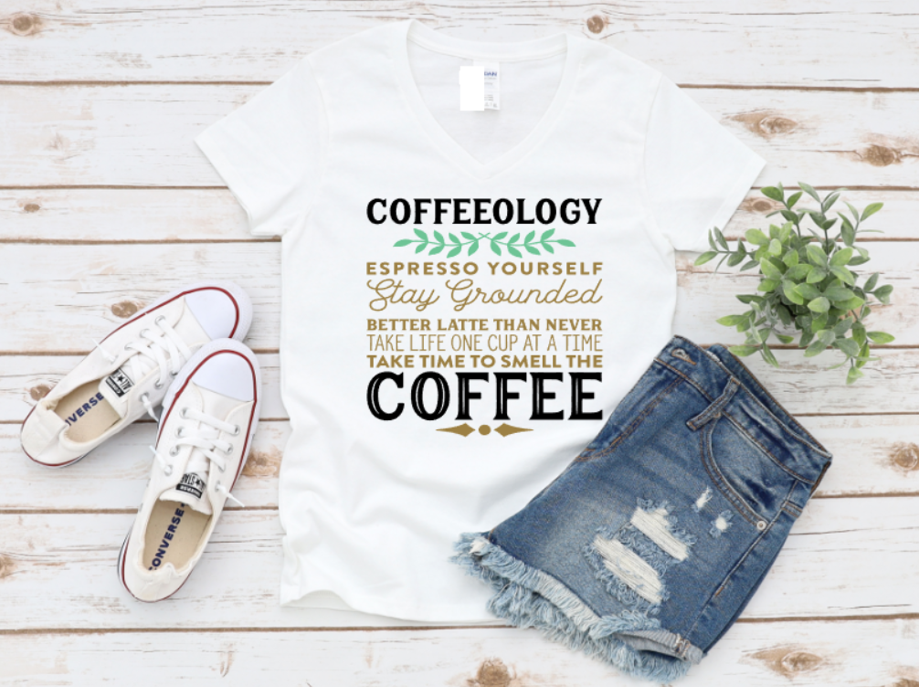White t-shirt with “Coffeeology” espresso-themed text in green, gold, and white, paired with distressed denim shorts and white Converse sneakers on a wooden background.