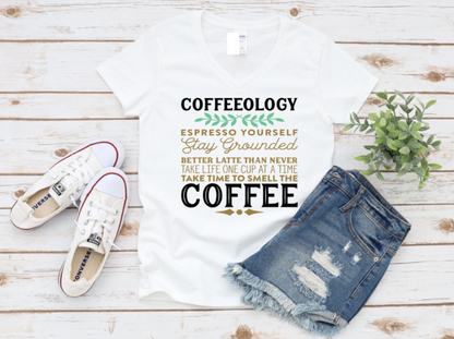 White t-shirt with “Coffeeology” espresso-themed text in green, gold, and white, paired with distressed denim shorts and white Converse sneakers on a wooden background.