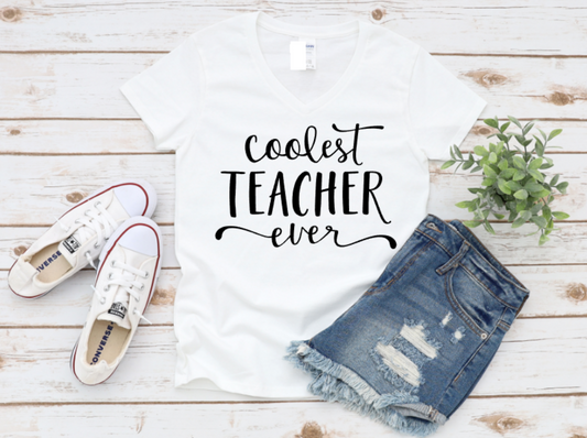 White tshirt with 'Coolest Teacher Ever' text, white sneakers, and denim shorts on a wooden surface.