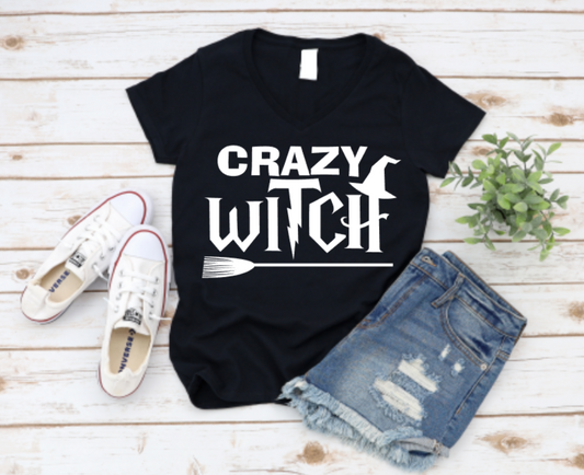 Black v-neck t-shirt with bold white "Crazy Witch" text featuring witch hat and broomstick graphic, styled with denim shorts and white Converse sneakers on a wood background.