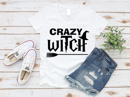 White v-neck t-shirt with bold white "Crazy Witch" text featuring witch hat and broomstick graphic, styled with denim shorts and white Converse sneakers on a wood background.