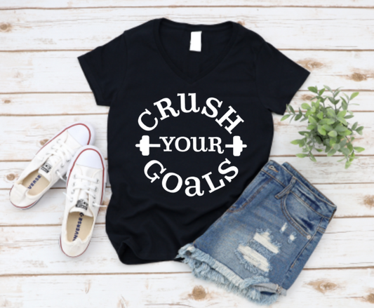 Flat lay of a black “Crush Your Goals” t-shirt with bold black lettering and dumbbell icons, styled with denim shorts, white Converse sneakers, and a plant on a wooden background.