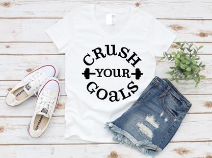 Flat lay of a white “Crush Your Goals” t-shirt with bold black lettering and dumbbell icons, styled with denim shorts, white Converse sneakers, and a plant on a wooden background.