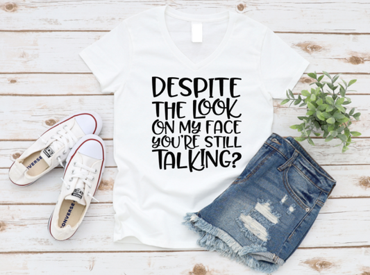 Flat lay of a white V-neck t-shirt with the sarcastic phrase “Despite the look on my face you’re still talking?” in white text, styled with ripped denim shorts, white Converse sneakers, and a small green plant on a wooden background.