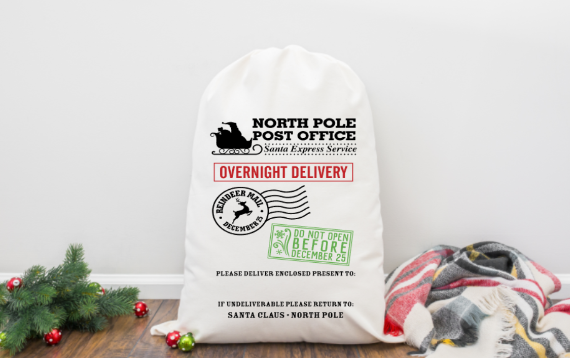 White Santa sack with festive text and graphics on a wooden surface with Christmas decorations.