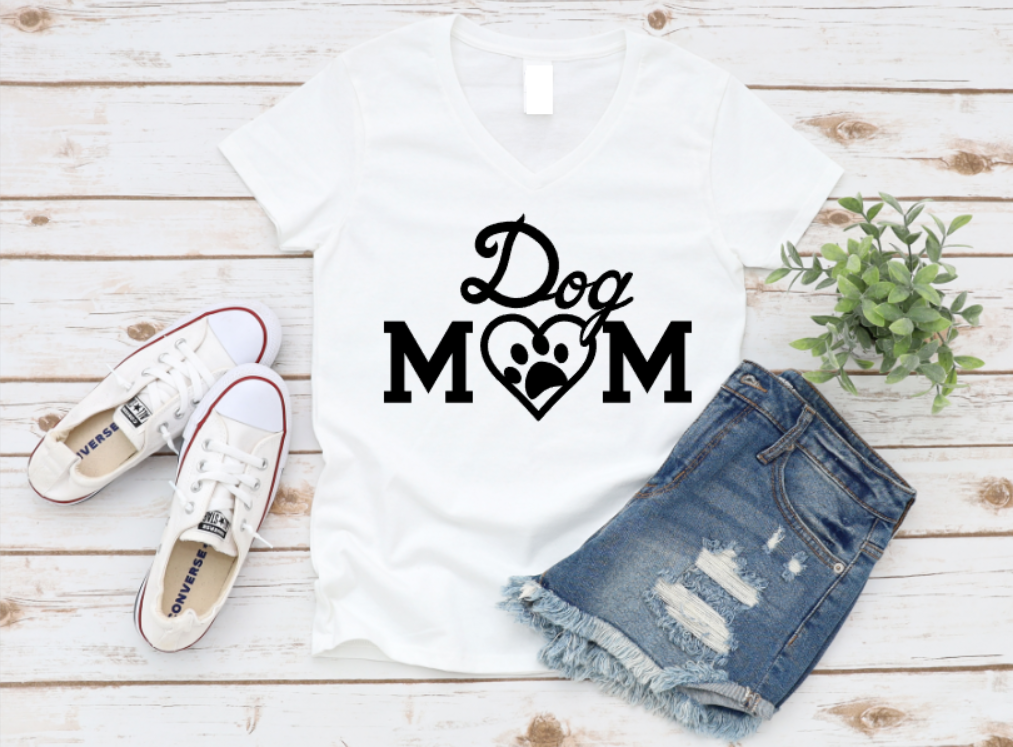 White t-shirt with “Dog Mom” text in black, featuring a heart-shaped paw print design, styled flat lay with denim shorts and white Converse sneakers on a wooden background.