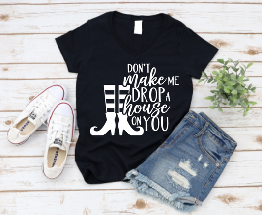 Flat lay of a black t-shirt with the phrase "Don't Make Me Drop a House on You" in white text, featuring witch legs and boots graphic. Styled with denim shorts and white Converse shoes on a rustic wood background.