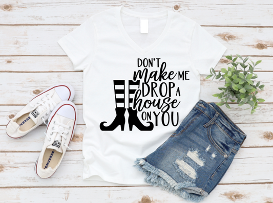 Flat lay of a white t-shirt with the phrase "Don't Make Me Drop a House on You" in white text, featuring witch legs and boots graphic. Styled with denim shorts and white Converse shoes on a rustic wood background.