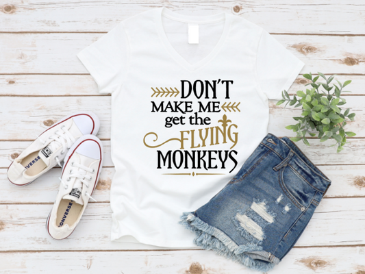 Flat lay of a white t-shirt with the phrase “Don’t Make Me Get the Flying Monkeys” in bold white and gold lettering, styled with ripped denim shorts, Converse sneakers, and a green plant on a wooden background.