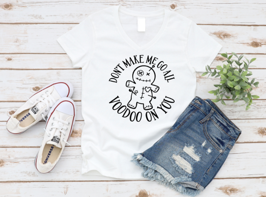 Flatlay of a white t-shirt with a white voodoo doll design and the phrase “Don’t Make Me Go All Voodoo On You,” styled with denim shorts and white sneakers on a wooden background.