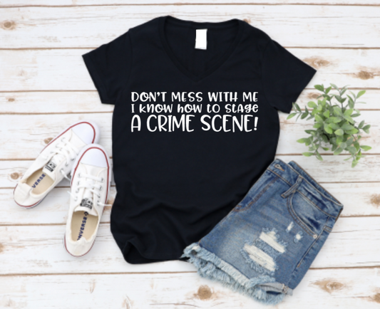Black v-neck t-shirt with the phrase “Don’t mess with me I know how to stage a crime scene” in bold white text, styled with denim shorts and white Converse sneakers on a rustic wood background.