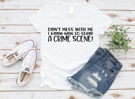 White v-neck t-shirt with the phrase “Don’t mess with me I know how to stage a crime scene” in bold white text, styled with denim shorts and white Converse sneakers on a rustic wood background.