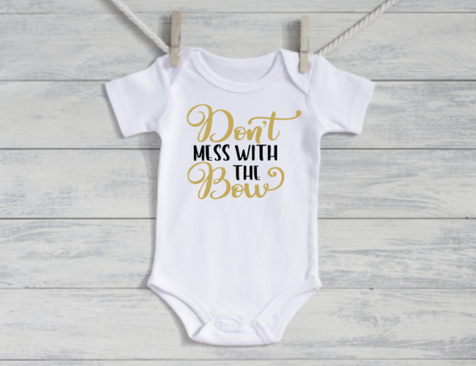White baby onesie with gold text 'Don't mess with the bow' on a wooden background